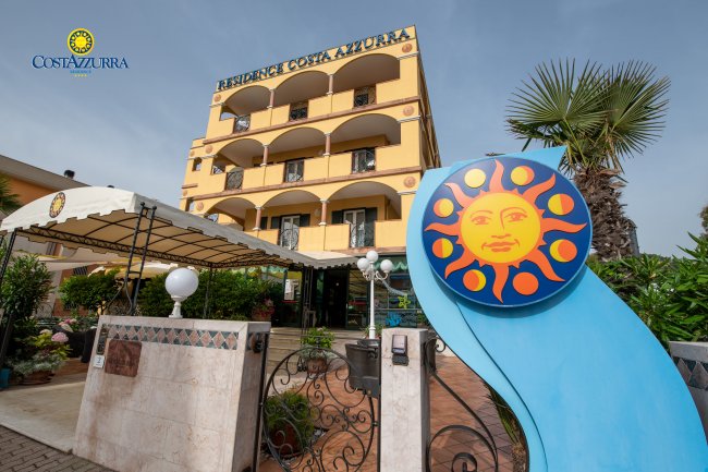 Esterno di un hotel giallo con insegna e giardino.