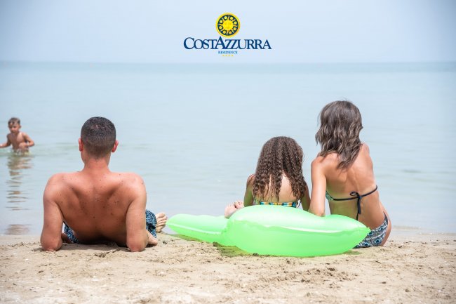 Tre persone in riva al mare, sedute sulla sabbia, con un gonfiabile verde.