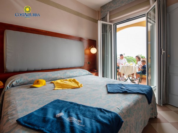 Camera d'hotel con letto matrimoniale e balcone con una famiglia al tavolo.