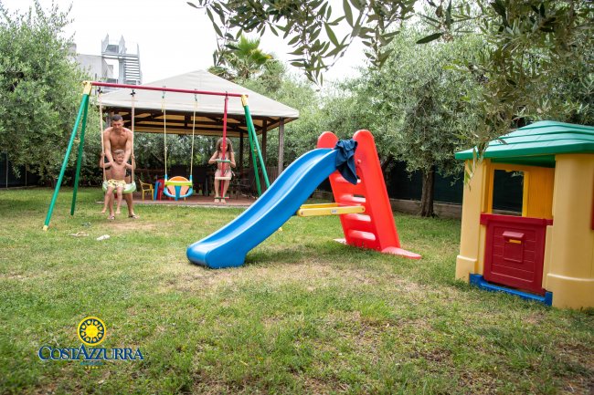 Area giochi con scivolo blu e altalene, circondata da alberi.