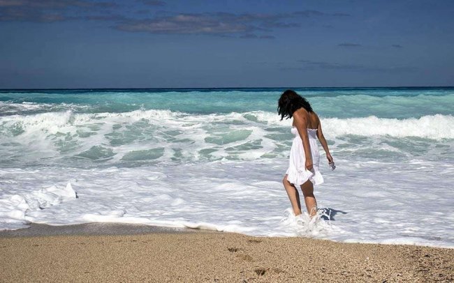 Una persona cammina sulla spiaggia con i piedi nell'acqua, indossa un abito bianco.