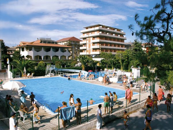 Piscina all'aperto con persone in costume da bagno, circondata da edifici residenziali e palme.