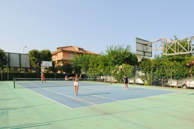 Due persone giocano a tennis su un campo all'aperto, circondato da alberi e edifici.