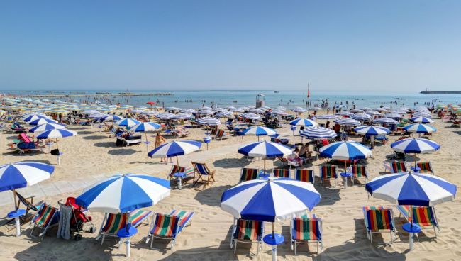 Spiaggia con ombrelloni blu e bianchi, lettini e bagnanti in mare.