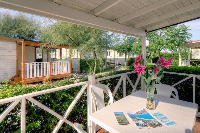 Una veranda con tavolo bianco e fiori, circondata da siepi e bungalow.