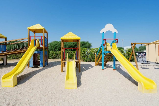 Gioco per bambini con tre scivoli gialli su una superficie sabbiosa.