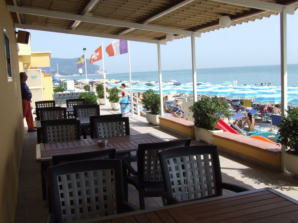 Terrazza con tavoli e sedie, vista su spiaggia con ombrelloni azzurri e mare.