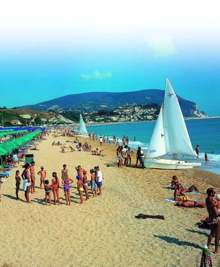 Spiaggia affollata con vele bianche e verdi, montagne sullo sfondo.