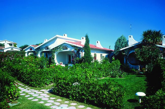 Bungalow bianco con giardino fiorito e vialetto, cielo azzurro.