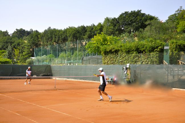 Due persone giocano a tennis su un campo in terra battuta, circondato da alberi.
