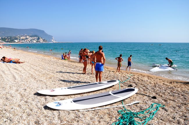 Spiaggia affollata con persone, paddleboard sulla sabbia e mare azzurro.