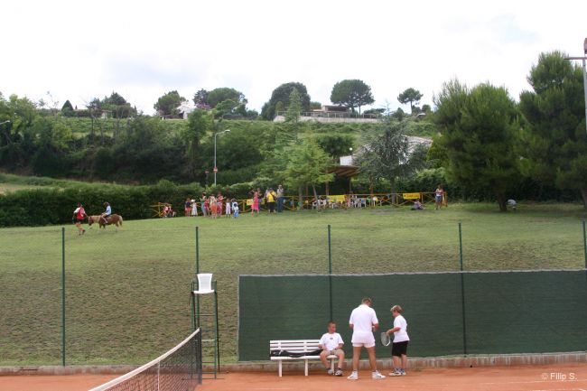 Campo da tennis con due persone, collina sullo sfondo con spettatori e cavalli.