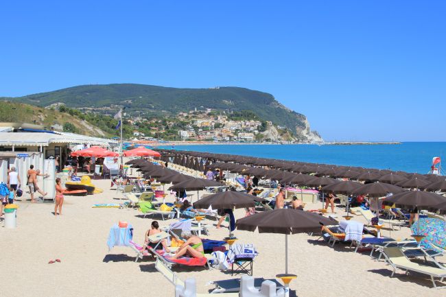 Spiaggia sabbiosa con ombrelloni scuri, mare blu e colline sullo sfondo.