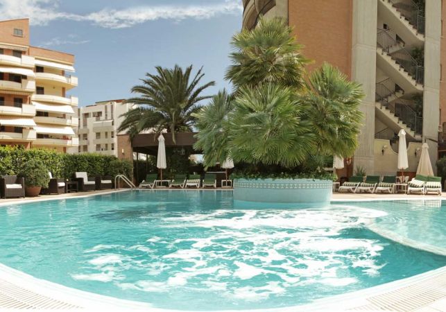 Piscina all'aperto con palme al centro, circondata da edifici e sedie a sdraio.