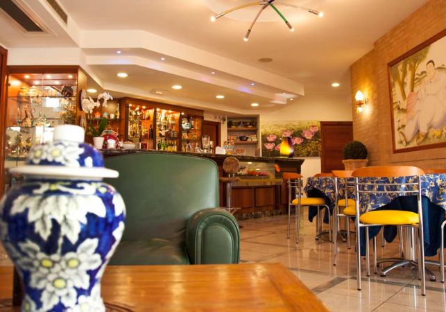 Interno di un bar con tavoli apparecchiati, divano verde e vaso decorato in primo piano.