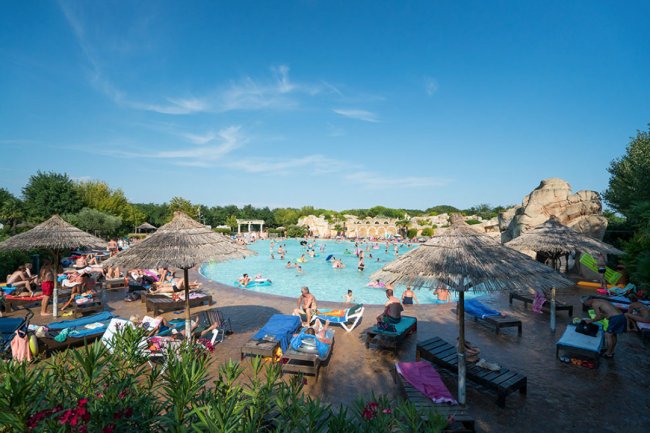 Spiaggia affollata con piscina, lettini e ombrelloni di paglia.