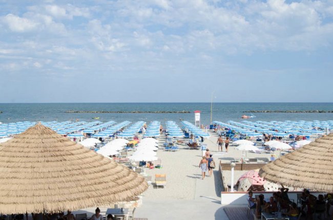 Spiaggia con ombrelloni azzurri e bianchi, mare sullo sfondo, cielo parzialmente nuvoloso.