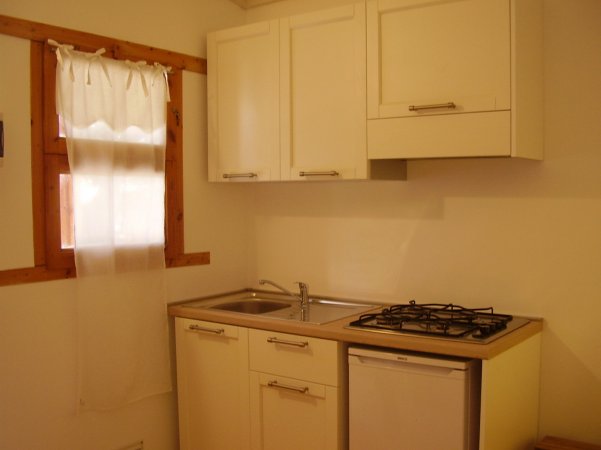 Cucina compatta con mobili bianchi, lavello, piano cottura e finestra con tende.