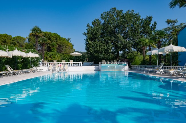Una piscina con lettini e ombrelloni, circondata da alberi verdi.