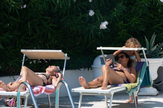 Due persone prendono il sole su lettini vicino a una piscina, con piante sullo sfondo.