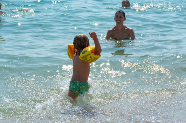 Bambino con braccioli arancioni entra nell'acqua, mentre un adulto lo osserva sorridendo.