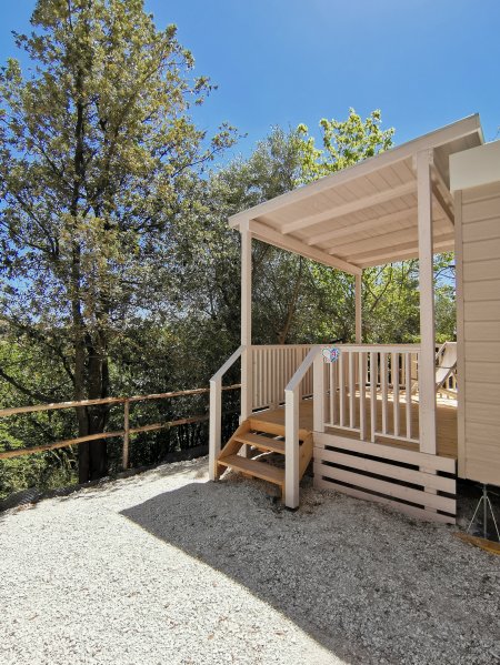 Una veranda in legno chiaro con scale, circondata da alberi, sotto un cielo azzurro.