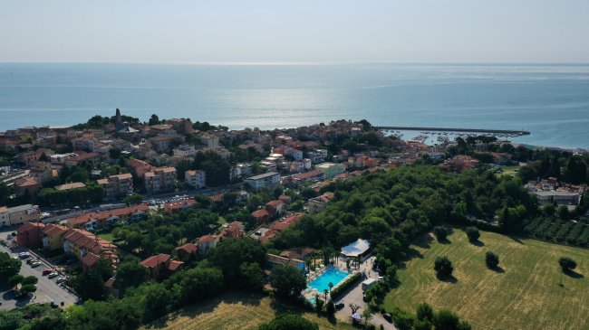 Vista aerea di un borgo costiero con edifici, campi e mare sullo sfondo.