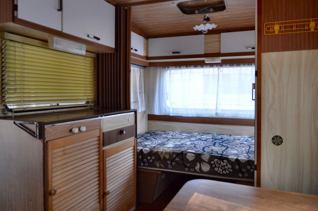 Interno di una roulotte vintage con letto, scaffali in legno e tende bianche.