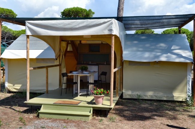 Una tenda con veranda arredata con tavolo e sedie, situata in un'area ombreggiata da alberi.