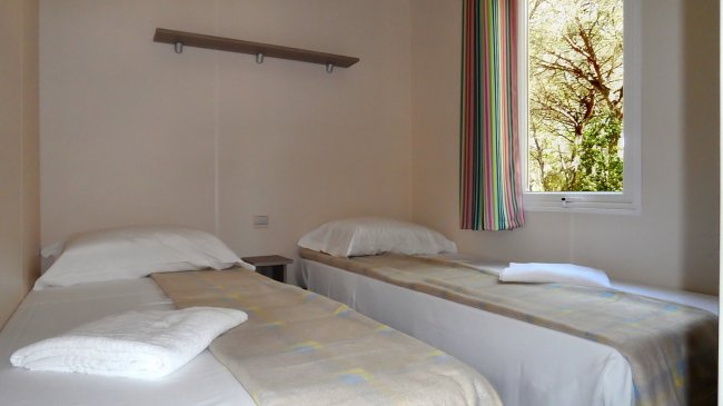 Due letti singoli con lenzuola, finestra con tende a righe e una mensola a muro.