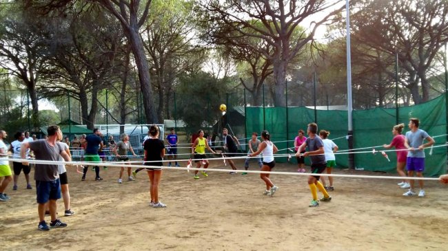Persone che giocano a pallavolo in un campo sabbioso, circondato da alberi.