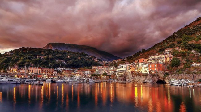 Un villaggio costiero riflesso in acqua, con cielo nuvoloso e montagne sullo sfondo.