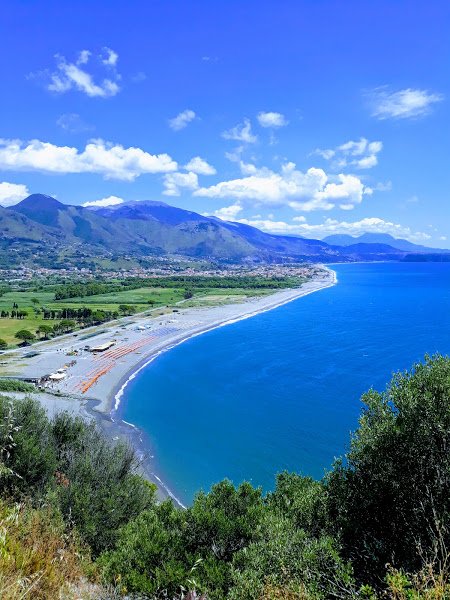 Vista aerea di una costa con spiaggia e mare blu, circondata da montagne e vegetazione.