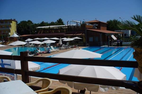 Una piscina con corsie e ombrelloni bianchi su un ponte, edificio in legno sullo sfondo.