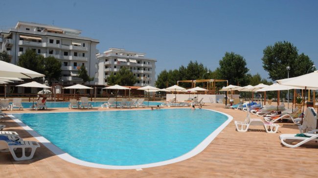 Una piscina con ombrelloni bianchi, lettini e edifici residenziali sullo sfondo.