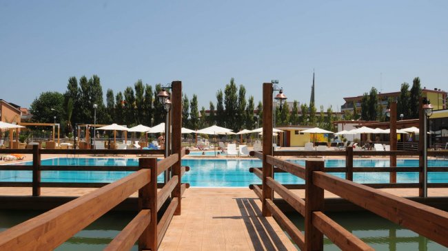 Pontile in legno sopra l'acqua conduce a una piscina, con ombrelloni e lettini sullo sfondo.