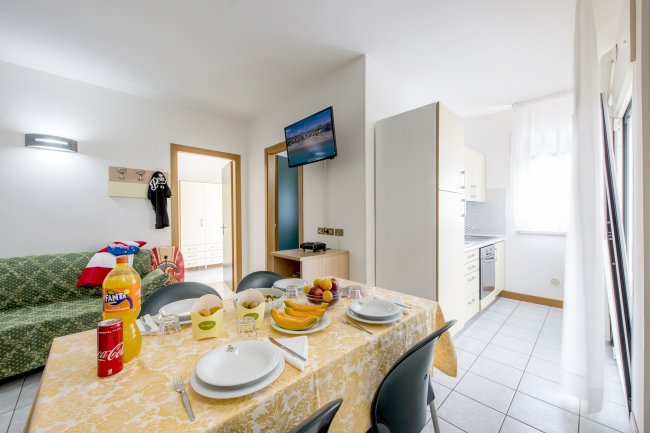 Appartamento con tavola da pranzo apparecchiata, divano verde, TV a parete e cucina sullo sfondo.