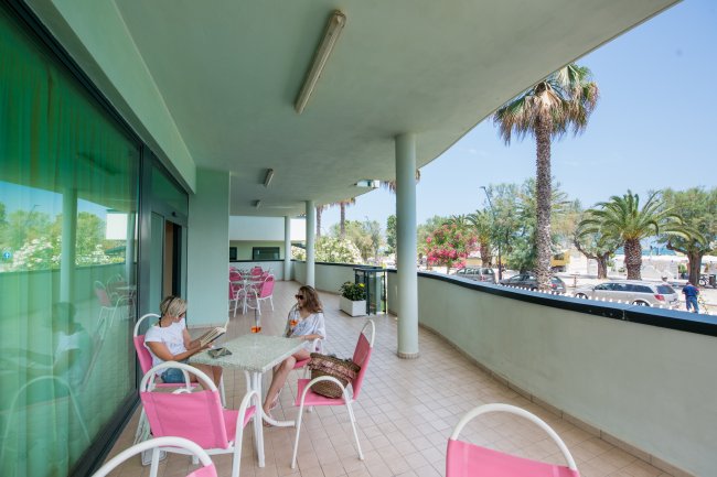 Due persone siedono su un ampio balcone con tavolo e sedie rosa, circondato da palme.