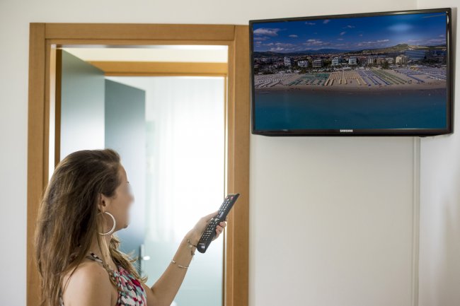 Una donna guarda un televisore a parete con vista aerea di una spiaggia, telecomando in mano.