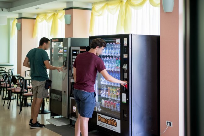 Due persone usano distributori automatici in una stanza luminosa con sedie e tavoli.