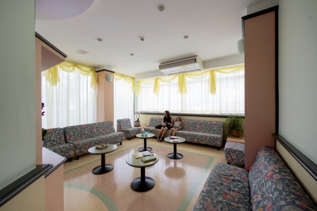 Sala d'attesa con divani colorati, tavolini e finestre luminose. Due persone sedute.