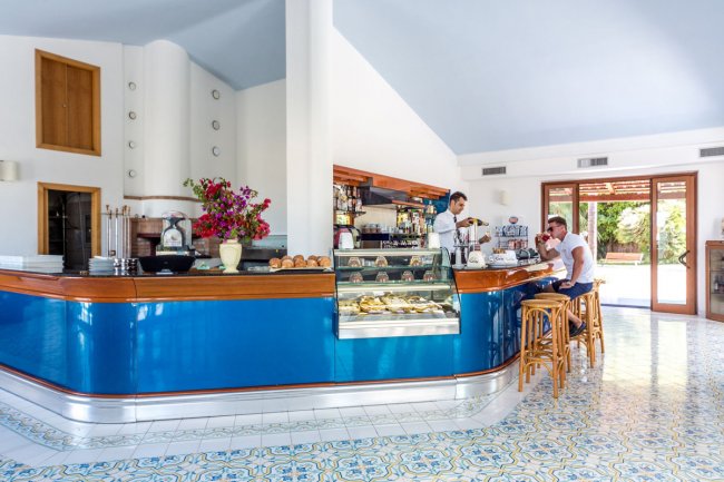 Interno di un bar con bancone blu, vetrina di dolci e due persone al bancone.