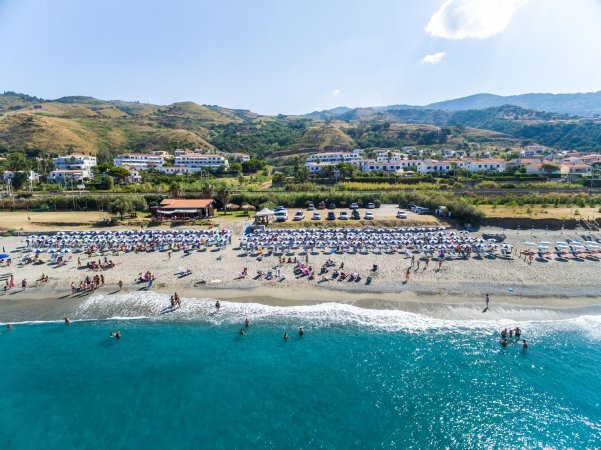 Spiaggia affollata con ombrelloni colorati, mare turchese e colline sullo sfondo.