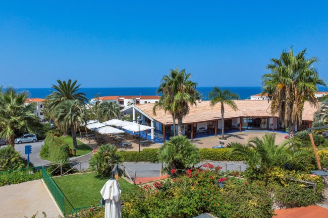 Un complesso turistico con palme, edifici bianchi e uno scorcio di mare blu in lontananza.
