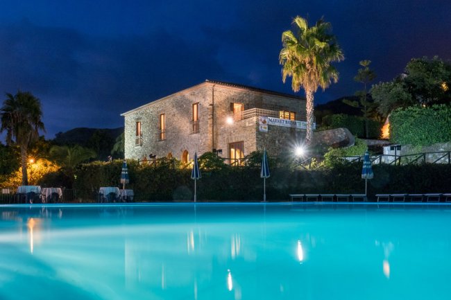 Edificio in pietra illuminato di notte, davanti a una piscina e palme sullo sfondo.