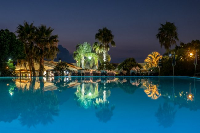 Piscina illuminata di notte circondata da palme e ombrelloni.