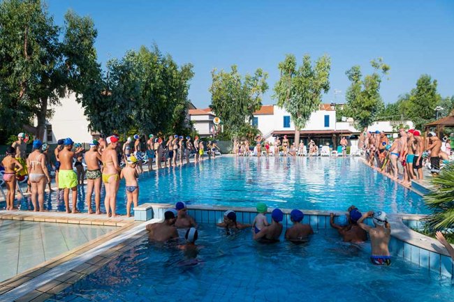 Piscina all'aperto con persone in fila, alcune in acqua, giornata di sole.