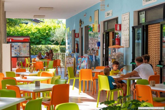 Una terrazza con tavoli e sedie colorate. Persone sedute e vegetazione visibile sullo sfondo.