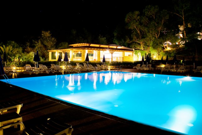 Piscina illuminata di notte con lettini attorno e vegetazione sullo sfondo.