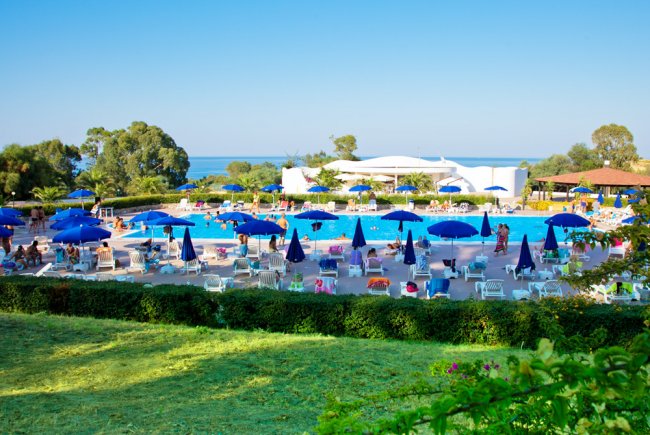 Ampia piscina con ombrelloni blu e lettini, vista mare e vegetazione circostante.